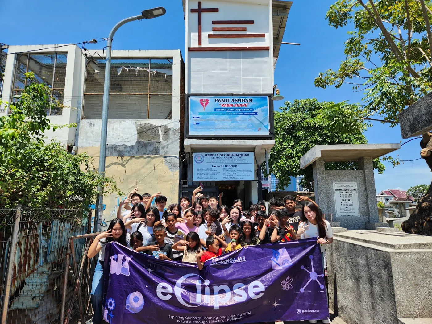 Eclipse volunteers and children holding a purple Eclipse banner outside Kasih Agape.